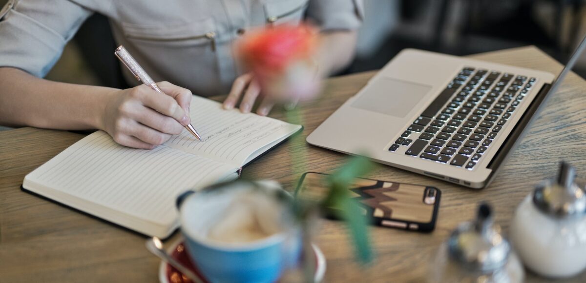 person writing on a notebook beside macbook