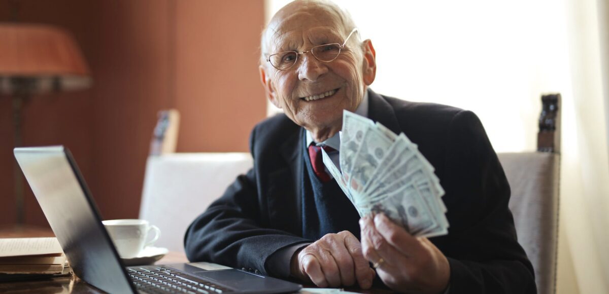 happy senior businessman holding money in hand while working on laptop at table