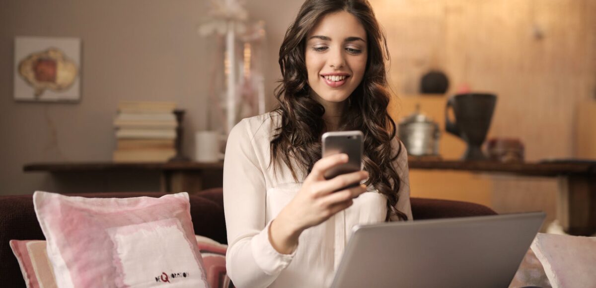 woman sitting on sofa while looking at phone with laptop on lap