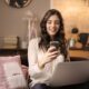 woman sitting on sofa while looking at phone with laptop on lap